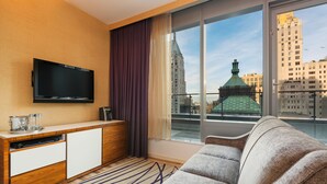 Ett modernt hotellrum med ett stort fönster, en platt-TV, en soffa, en träkonsol och utsikt över stadens byggnader.