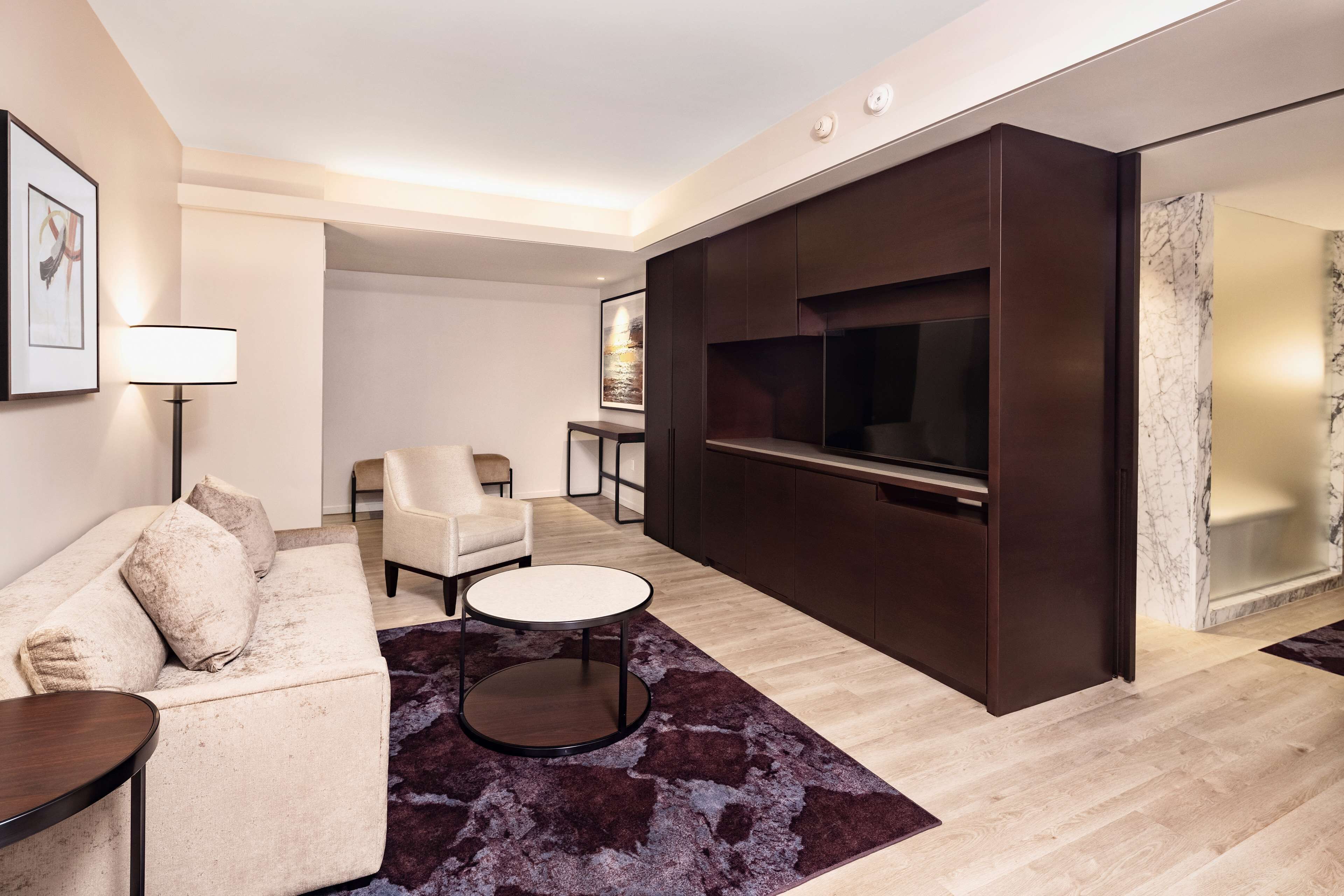A modern living room with a sofa, armchair, and round coffee table. A flat-screen TV is mounted on a dark wooden wall.