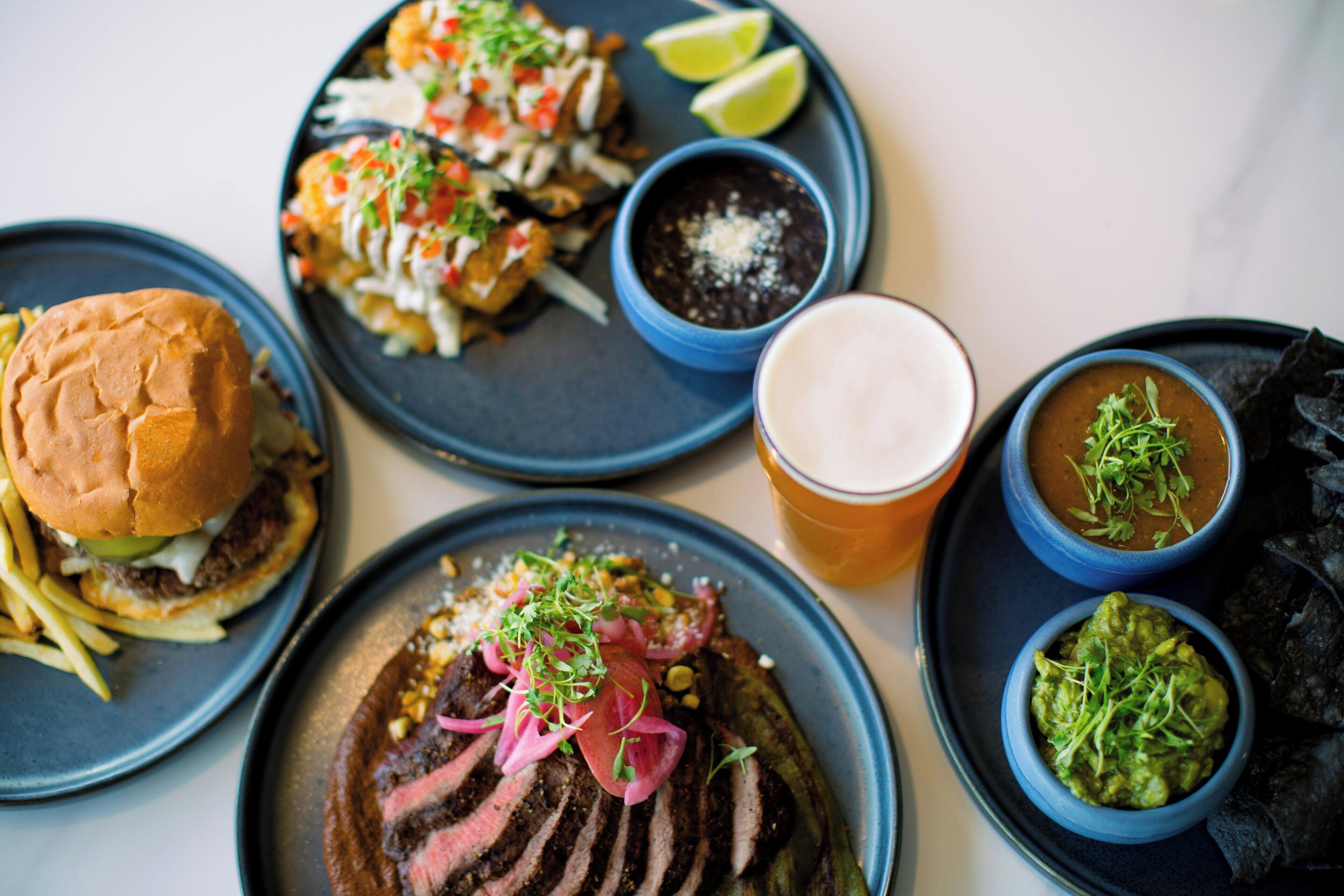 A variety of dishes including a burger, steak, and bowls of guacamole and salsa, served on blue plates.