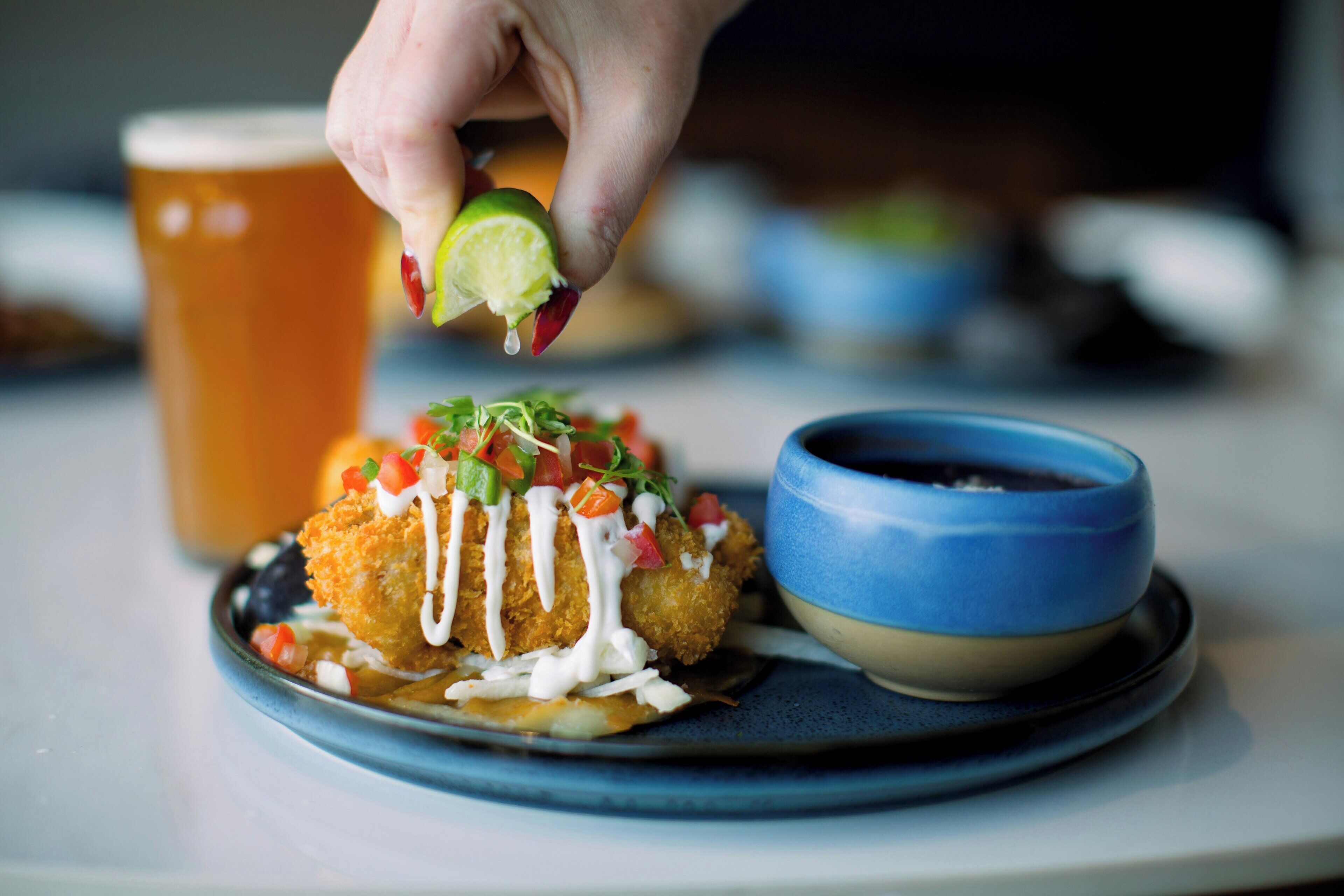 A hand holding a lime slice above a dish with a creamy sauce, garnished with diced vegetables, on a plate with a blue rim.