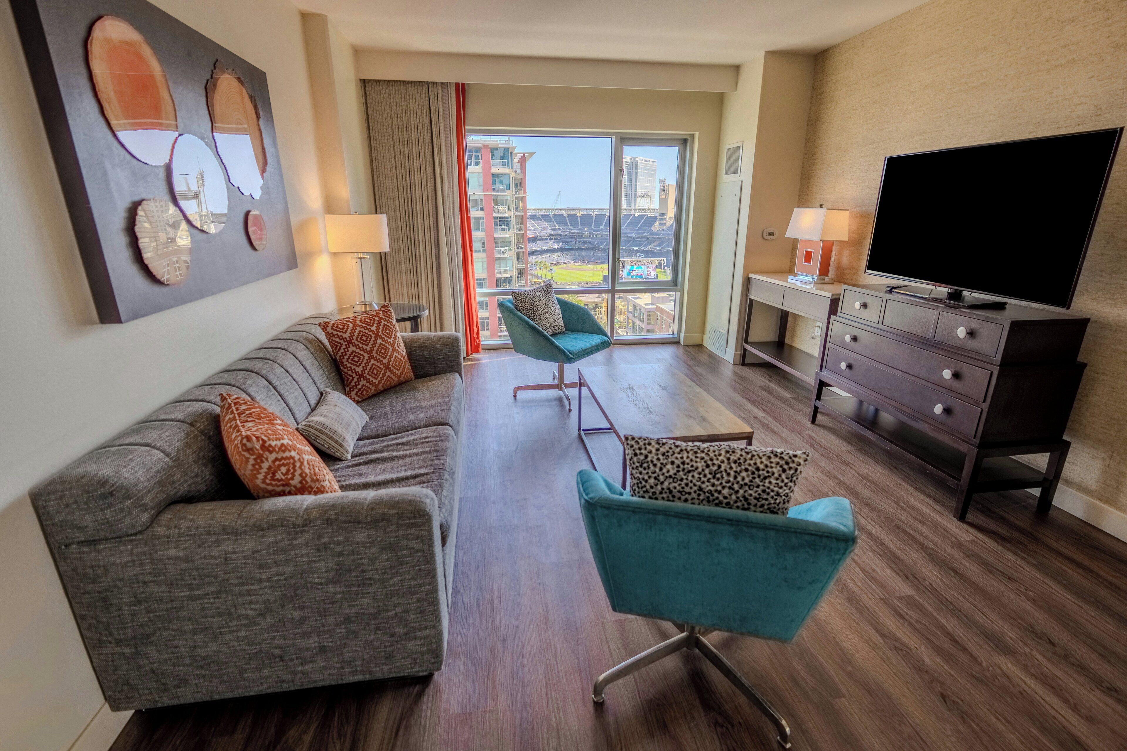 A hotel room with a sofa, two armchairs, a TV, a desk, and a large window with a view of a stadium.