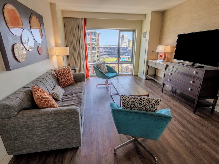 A hotel room with a sofa, two armchairs, a TV, a desk, and a large window with a view of a stadium.