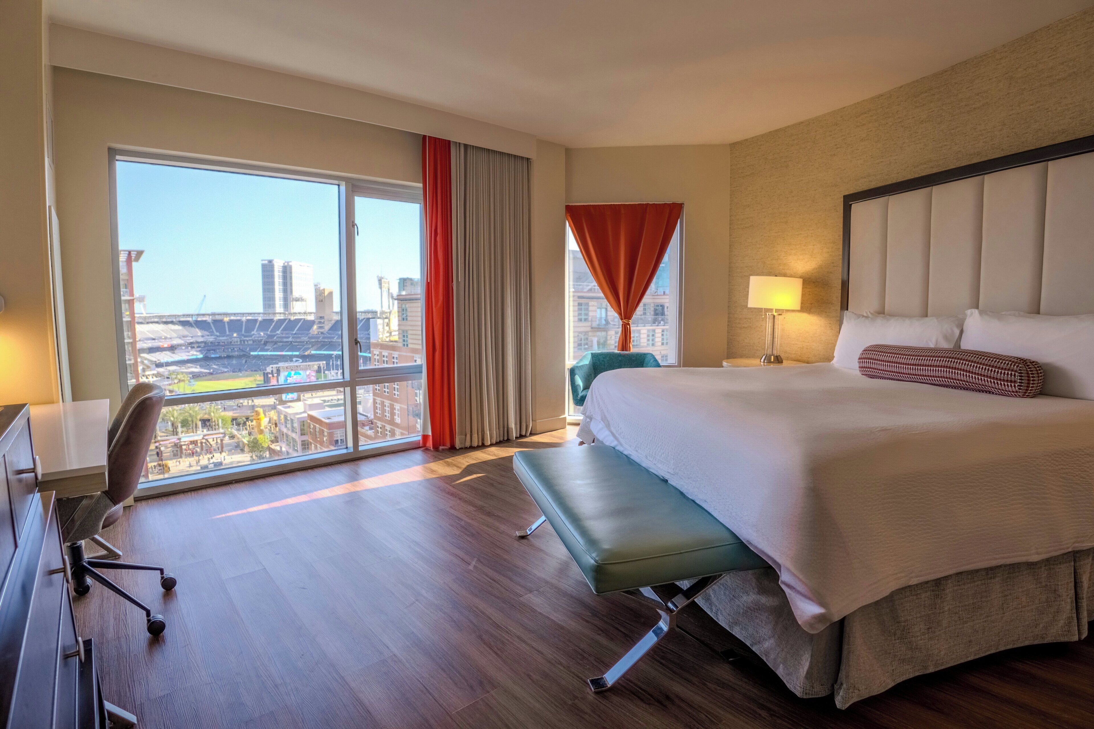 Habitación de hotel con una cama grande, un escritorio, una silla, un banco y vistas a un paisaje urbano a través de un ventanal amplio.
