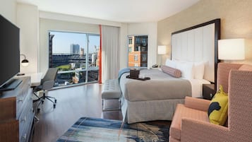 A hotel room with a large bed, a desk with a chair, a television, and a view of a cityscape through the window.