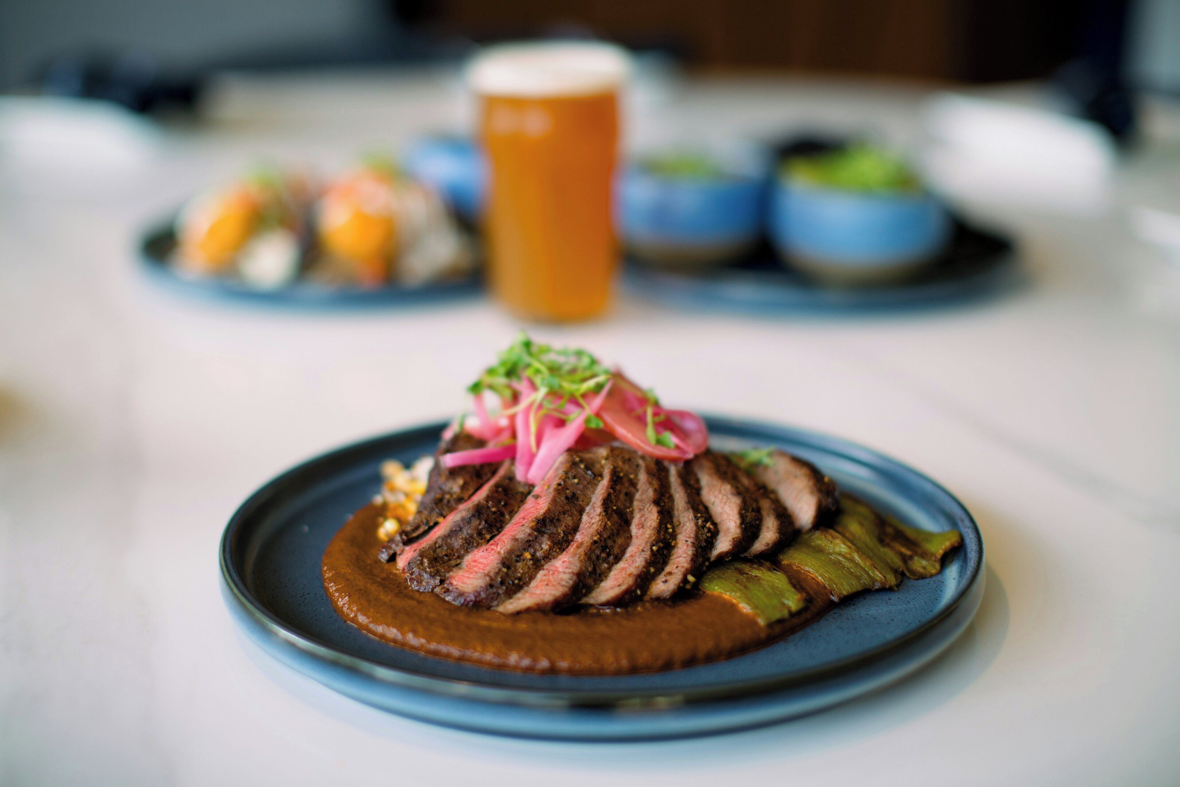 A plate of sliced steak with a sauce and garnish, a glass of beer, and other dishes in the background.