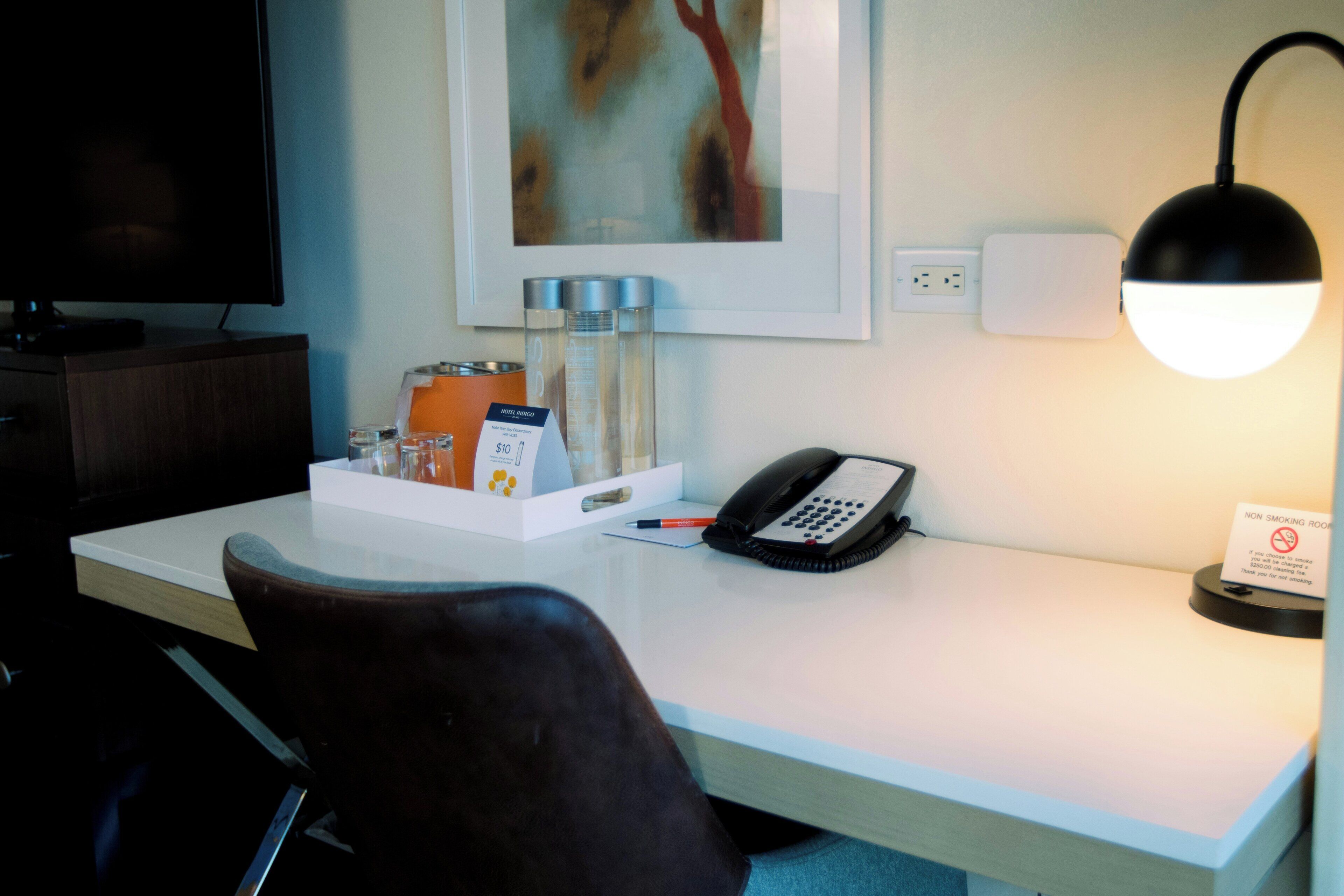 A hotel room desk with a telephone, a non-smoking sign, a framed artwork, and a small table with toiletries.