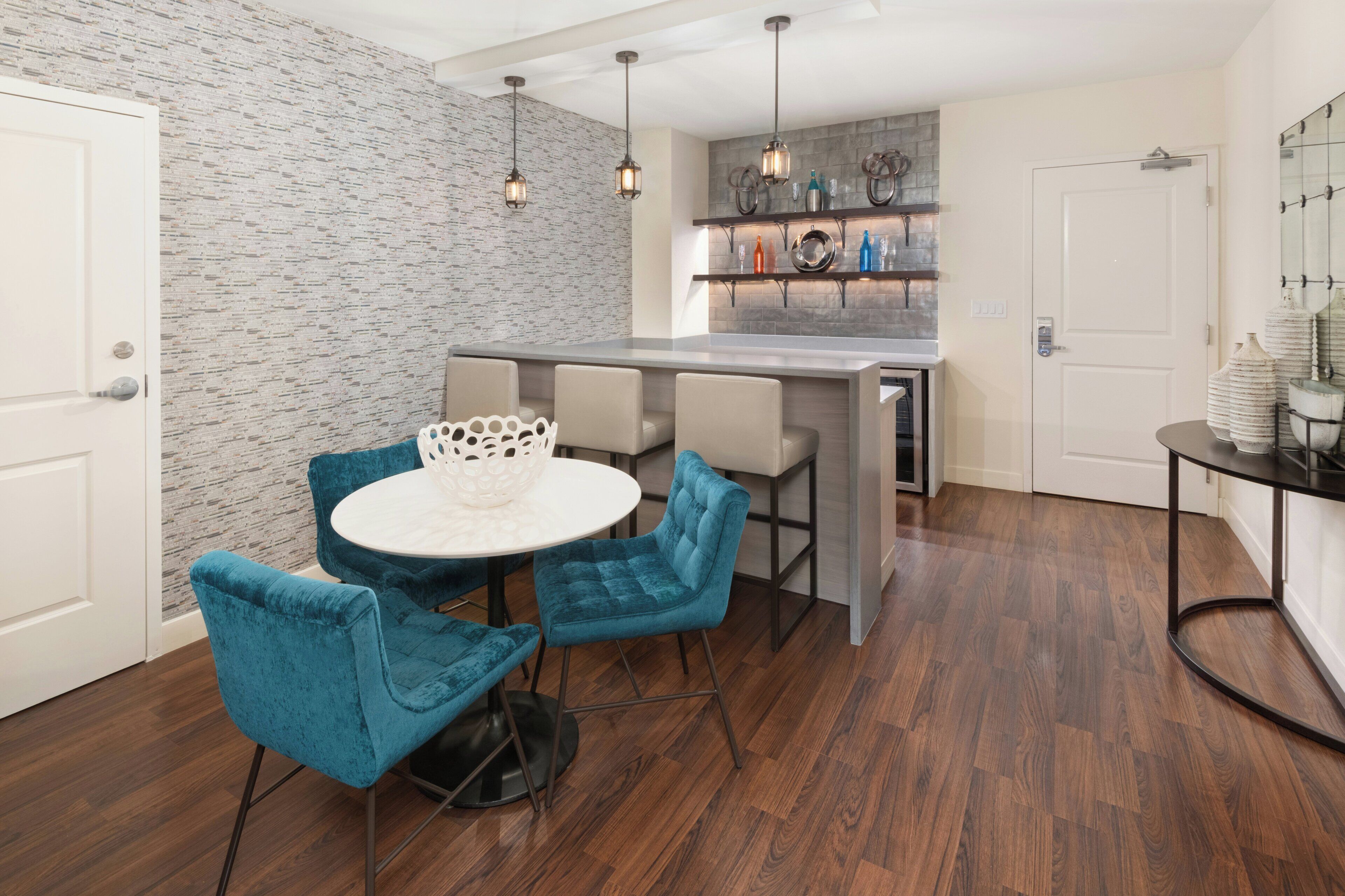 Una cocina moderna con una isla integrada, una zona de comedor con una mesa redonda y sillas azules, y una pared con estampado de ladrillo.