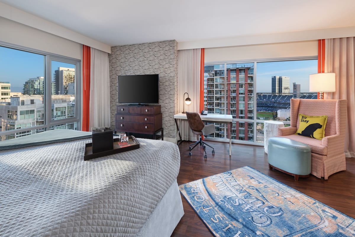 Une chambre d’hôtel avec un grand lit, une télévision, un bureau et une chaise. Les fenêtres offrent une vue sur le paysage urbain.