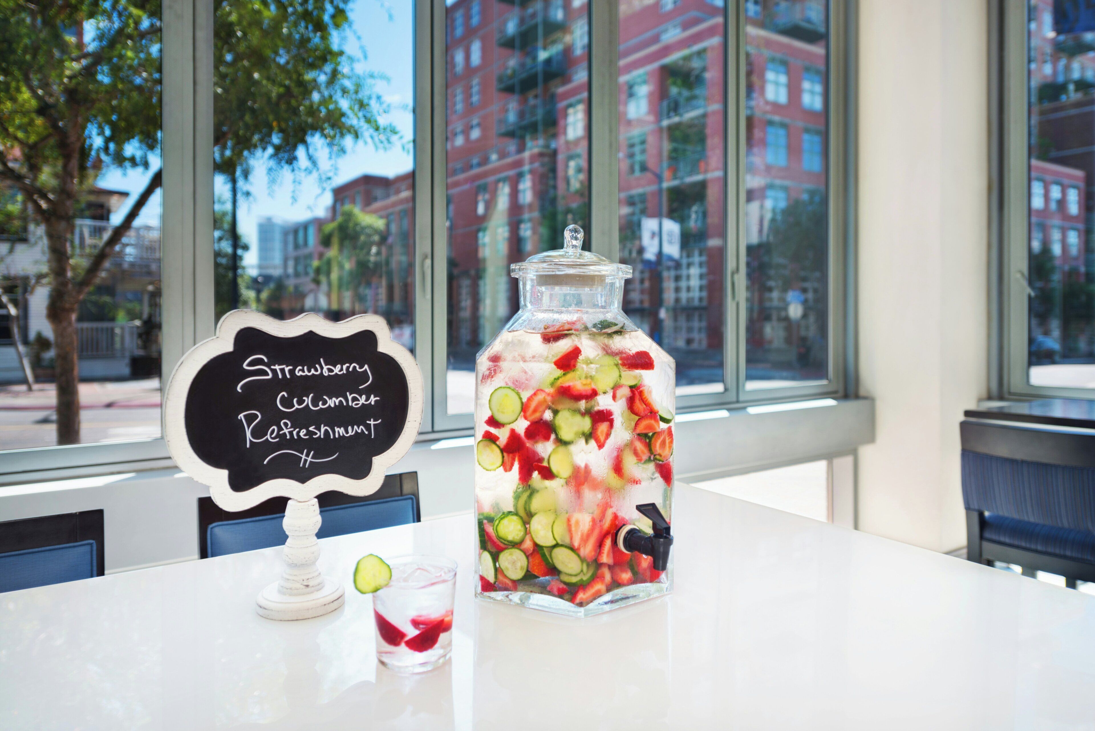 A clear glass jar filled with strawberries, cucumbers, and a straw, placed on a table with a chalkboard sign reading 'Strawberry Cucumber Refreshment.'