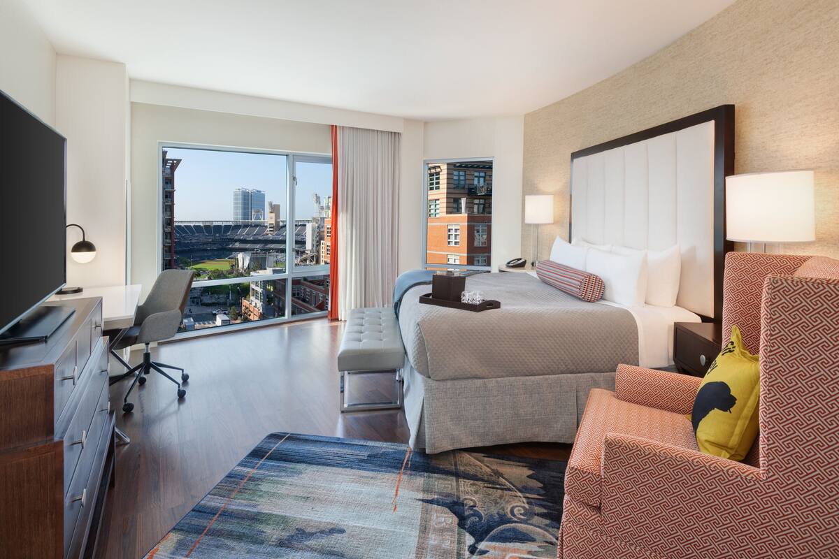 Une chambre d’hôtel avec un grand lit, un bureau avec une chaise, une télévision et une vue sur le paysage urbain par une grande fenêtre.