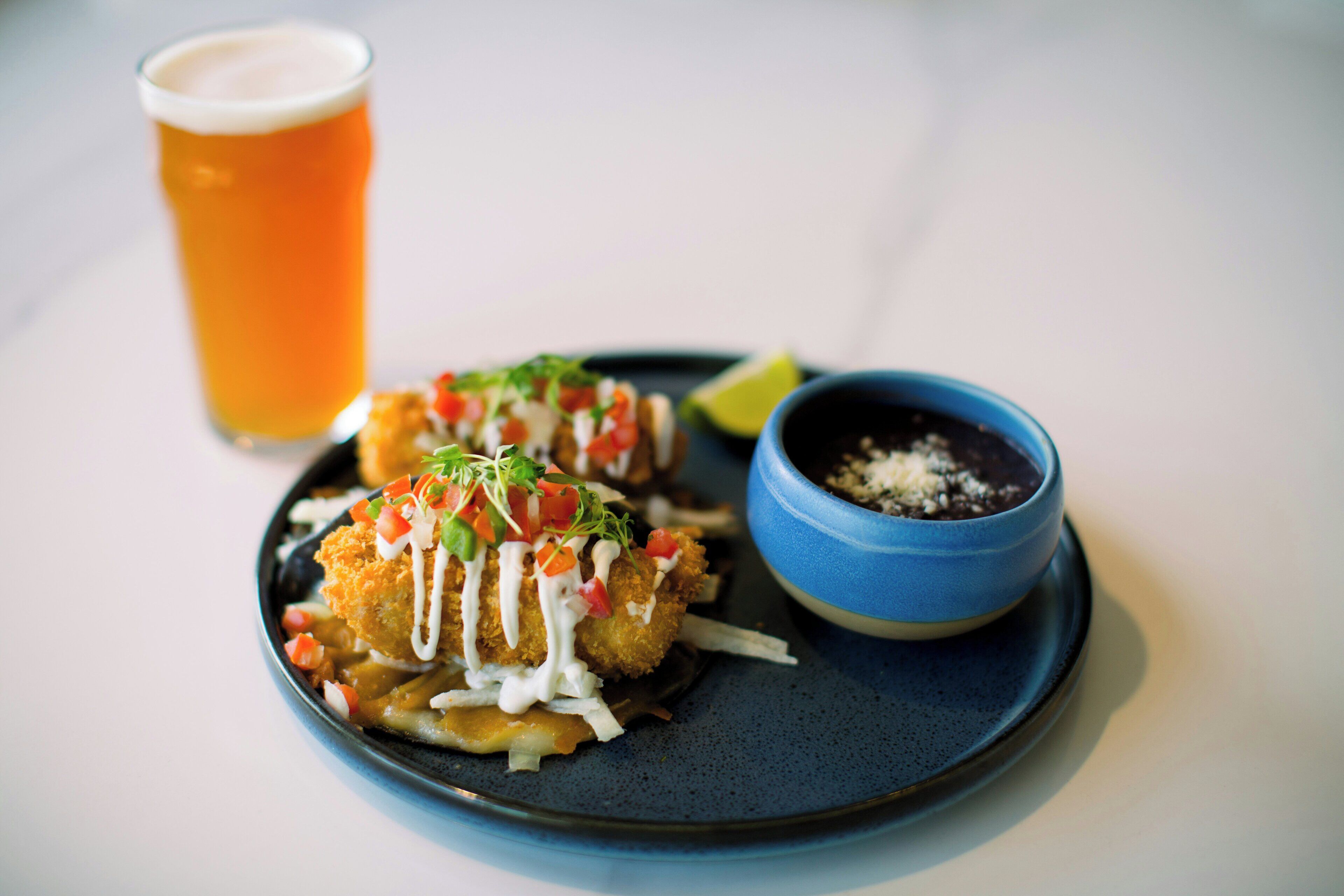 A plate with two fried items topped with sauce and vegetables, a glass of beer, and a bowl of sauce.