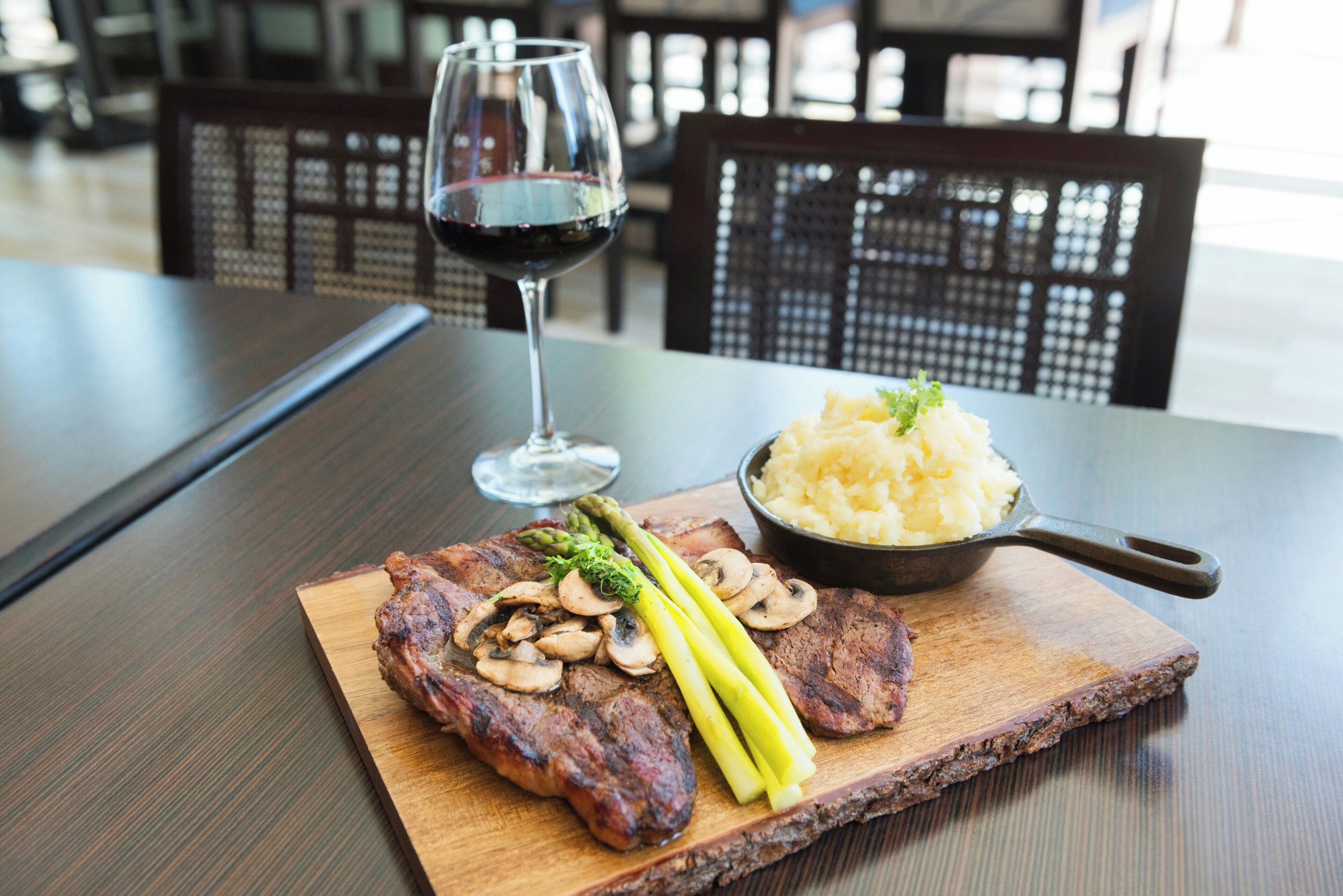 A wooden cutting board with steak, mushrooms, asparagus, and a bowl of mashed potatoes on a table.