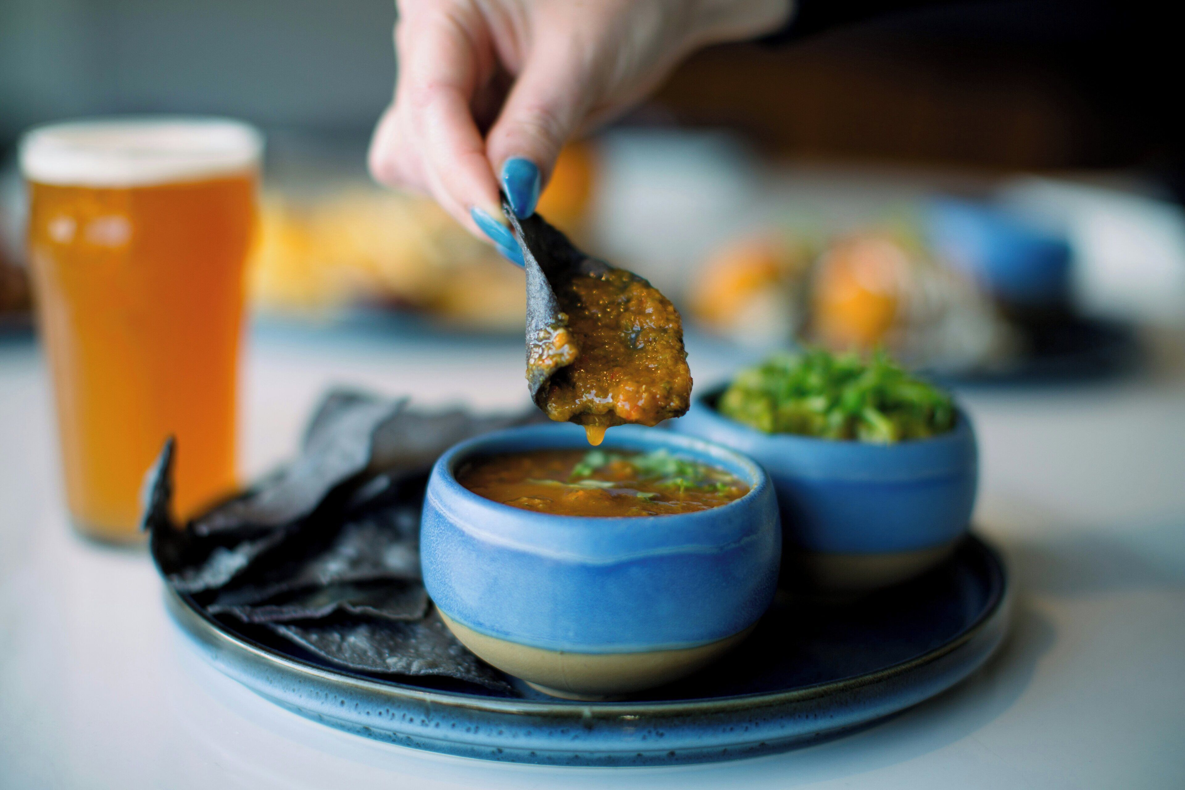A hand dipping a food item into a blue bowl, with a glass of beer and another bowl of food in the background.