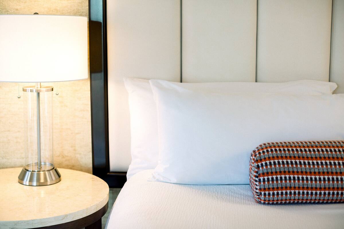 Un lit bien fait, avec du linge de lit blanc et un coussin à motifs, une table ronde avec une lampe, et un mur orné d’un panneau décoratif vertical.