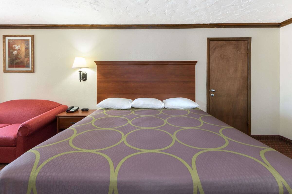A hotel room with a bed, a nightstand, a red chair, a wooden door, and a wall-mounted lamp.