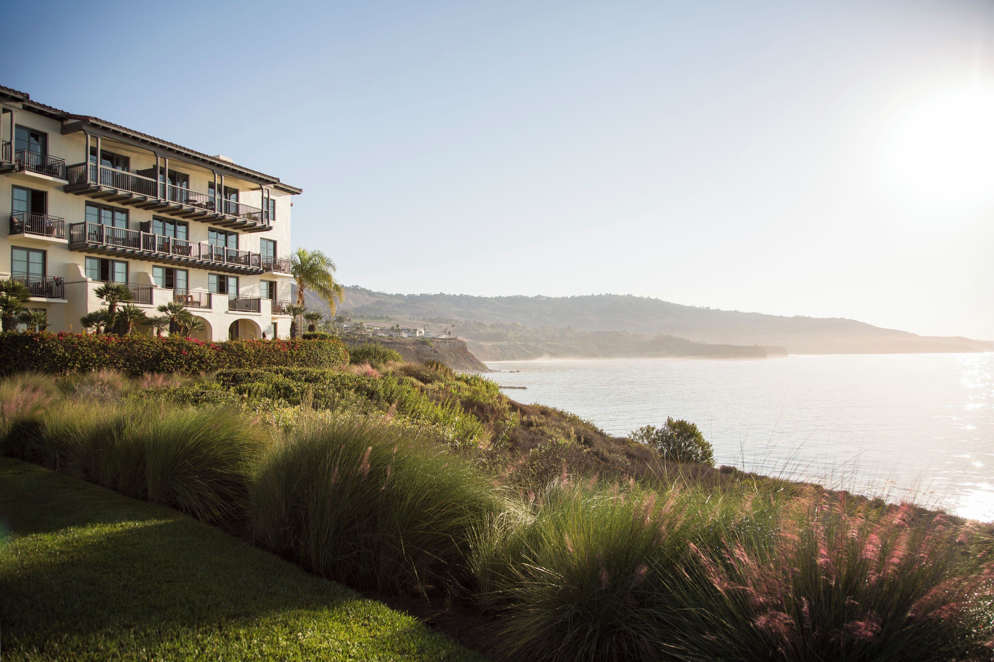 Ein Hotel an der Küste mit mehreren Balkonen, einem angelegten Garten und Blick auf das Meer und die entfernten Hügel.