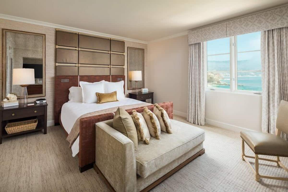 A hotel room with a large bed, a sofa, a chair, a nightstand, and a view of the sea.