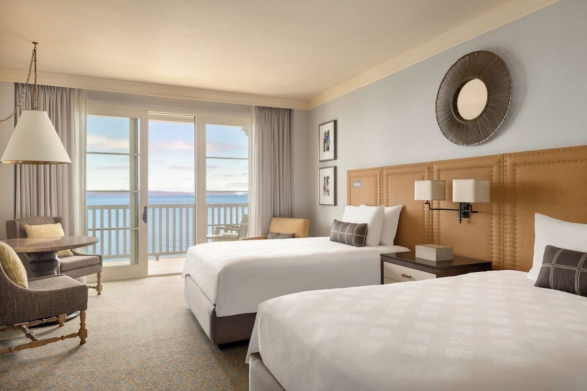 Habitación de hotel con dos camas, zona de estar con una silla y una mesa, ventana grande con vista al mar y un espejo decorativo en la pared.