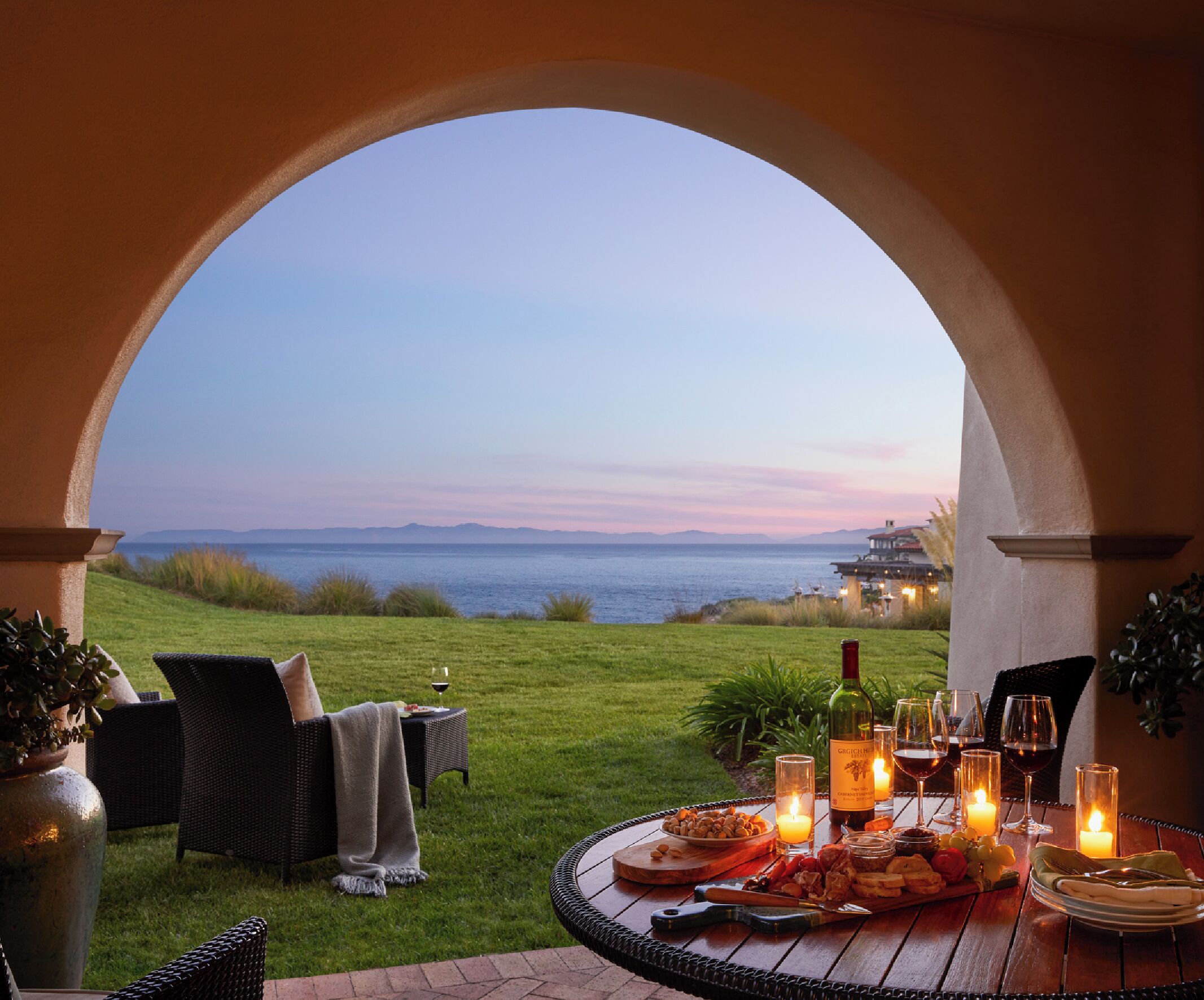 An outdoor dining area with a view of the sea, a table set with food and wine, and a cozy seating arrangement.