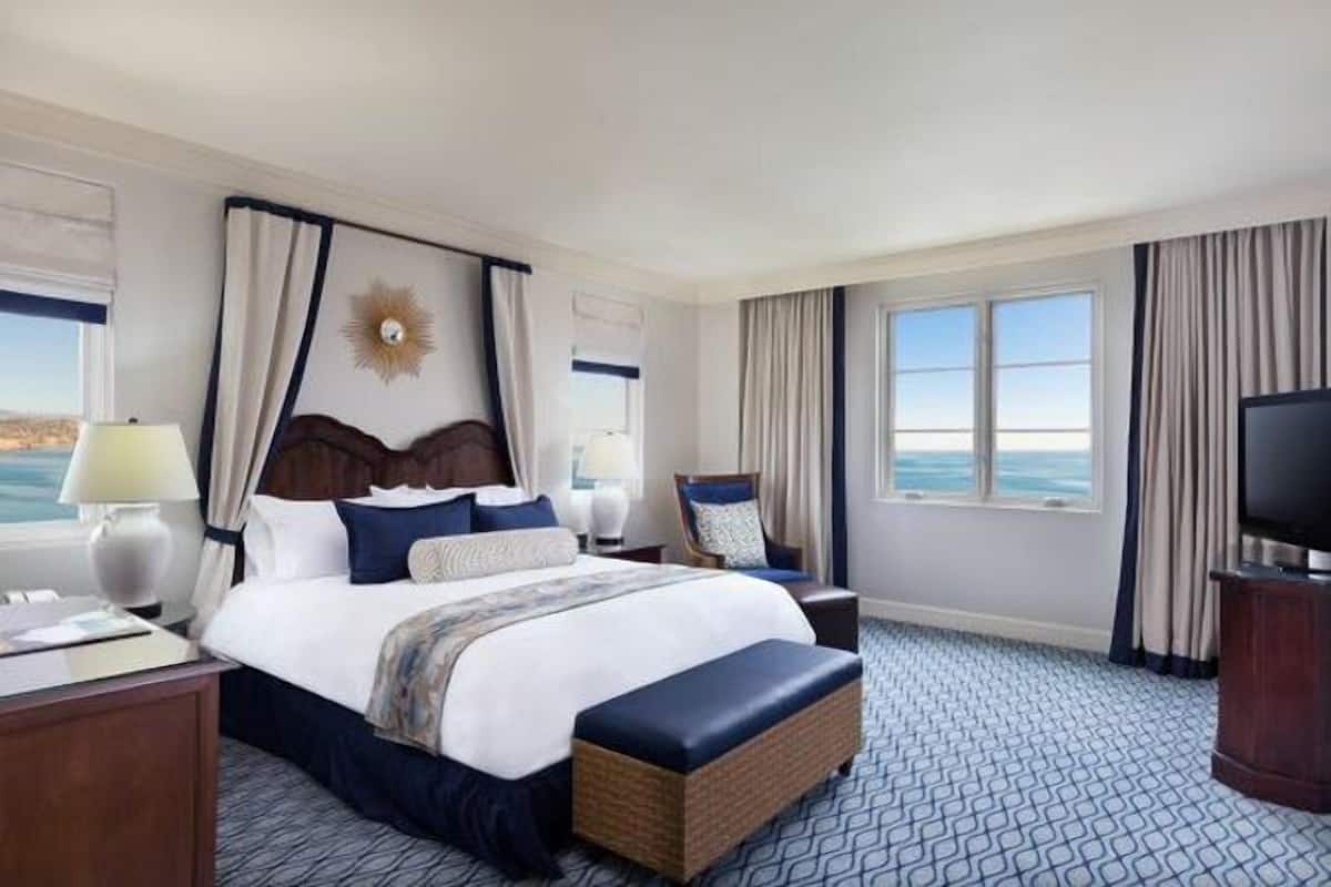 Una habitación de hotel con una cama grande, televisión, mesita de noche, lámpara y vista al océano.