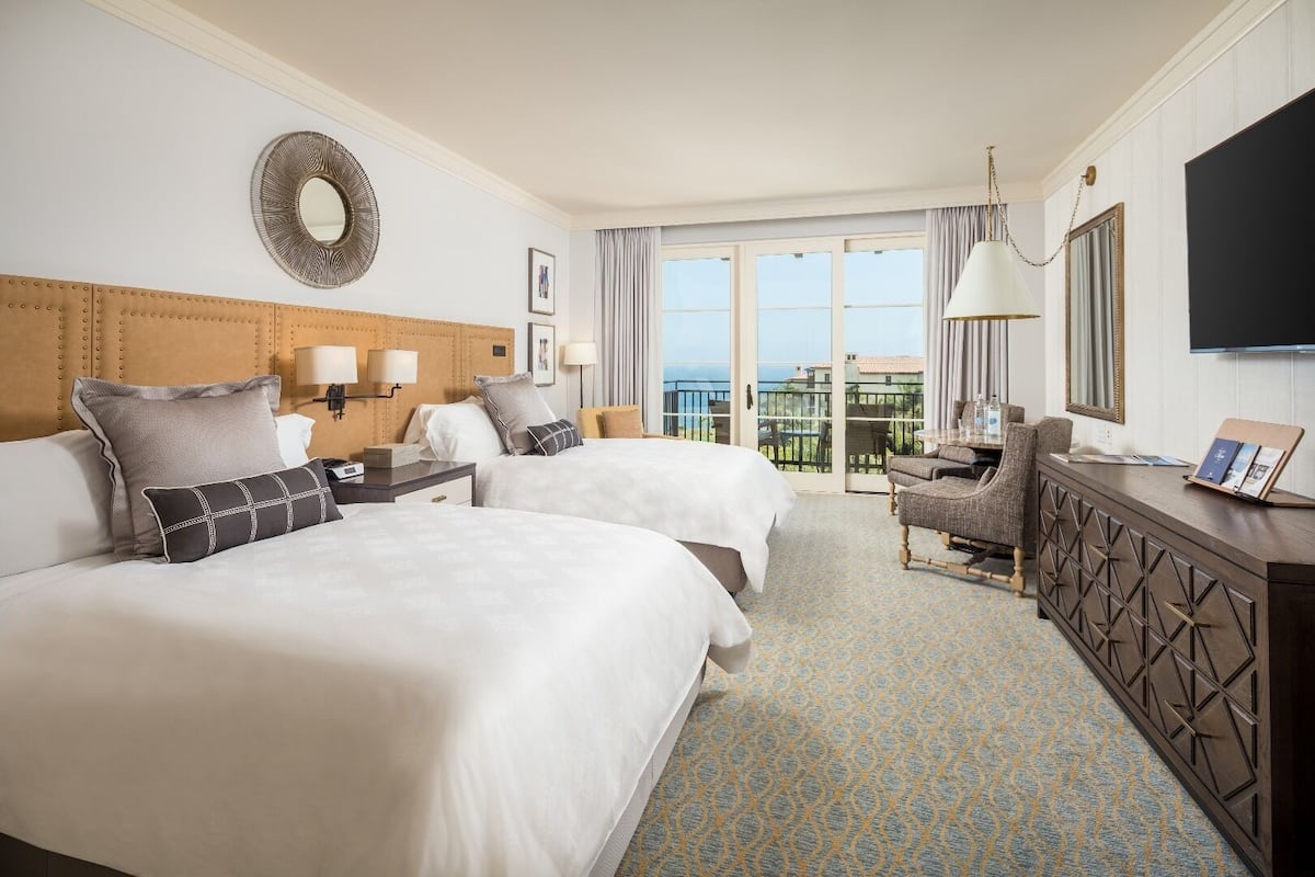 Habitación de hotel con dos camas, un escritorio, una silla, un televisor y un balcón con vistas.