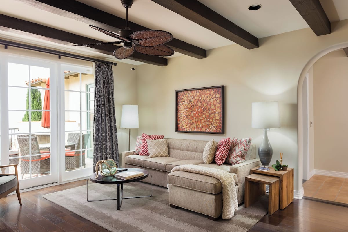 A living room with a sofa, coffee table, and lamps. There is a ceiling fan and a large window with curtains.