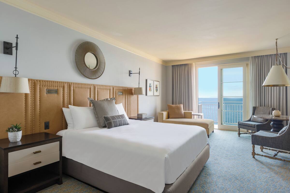 Habitación de hotel con una cama grande, una zona de estar con dos sillones, una mesita y una planta en maceta, además de vistas al océano a través de la ventana.