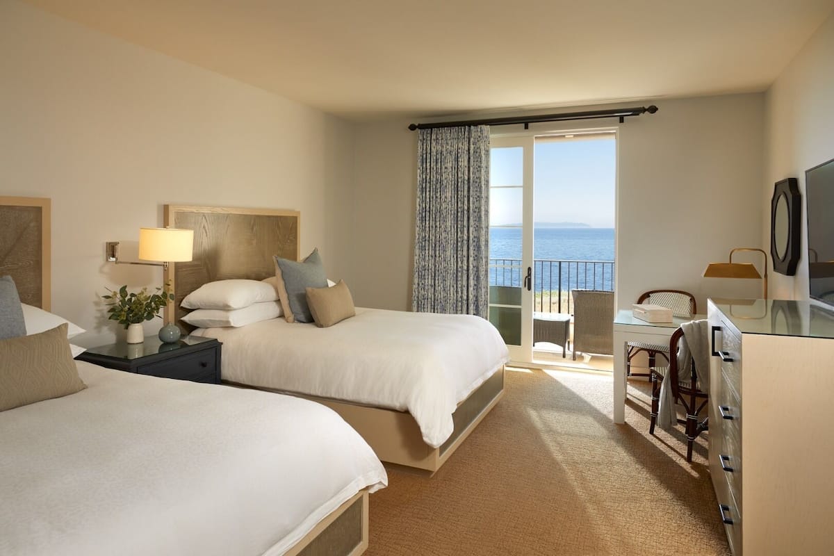 A hotel room with two beds, a desk, a chair, a TV, and a balcony with a view of the sea.