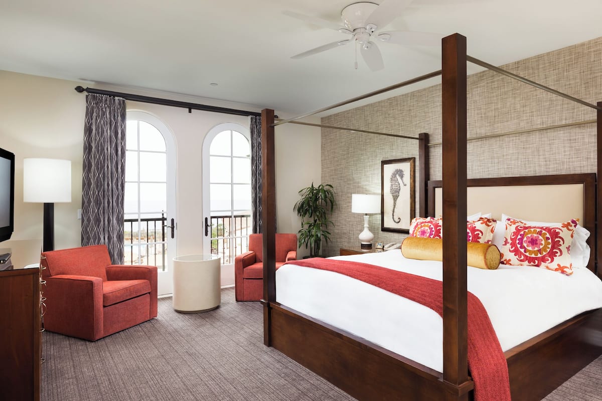 A four-poster bed with a red throw blanket, two red armchairs, a round table, a TV, and a balcony with curtains.