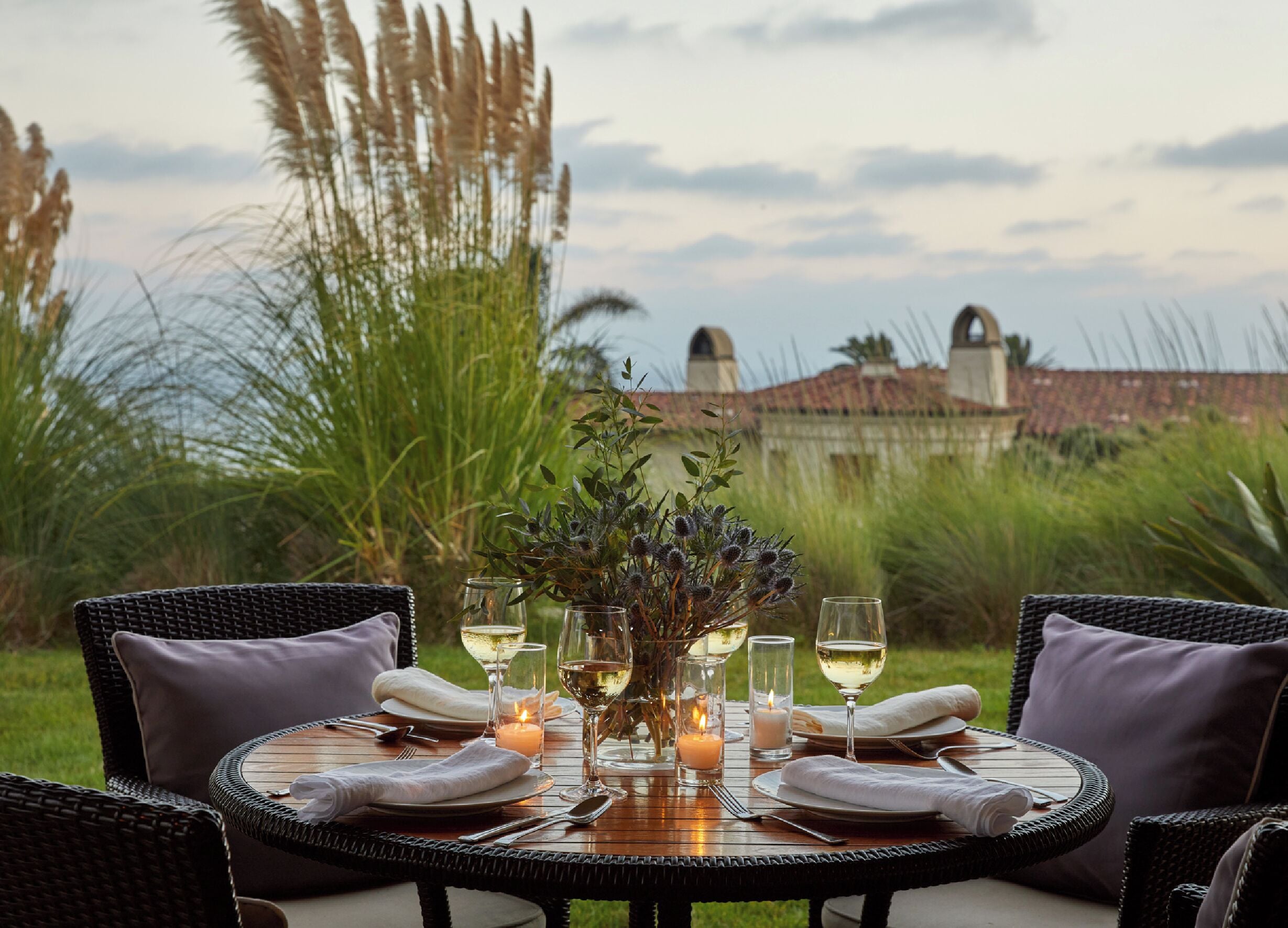 A round dining table set for four with white napkins, wine glasses, and candles, surrounded by wicker chairs with cushions, set against a backdrop of tall grasses and a building.