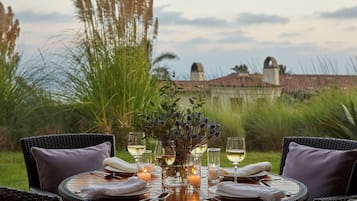 A round dining table set for four with white napkins, wine glasses, and candles, surrounded by wicker chairs with cushions, set against a backdrop of tall grasses and a building.