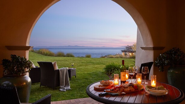 Un área de comedor al aire libre con vistas al mar, una mesa dispuesta con comida y vino, y una zona de estar acogedora.