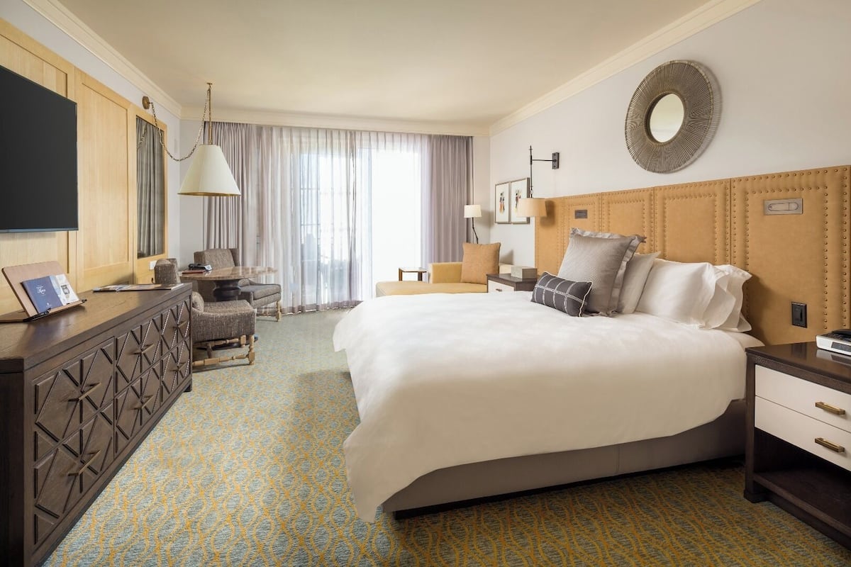 A hotel room with a large bed, a desk with a laptop, a chair, a sofa, and a TV on the wall.