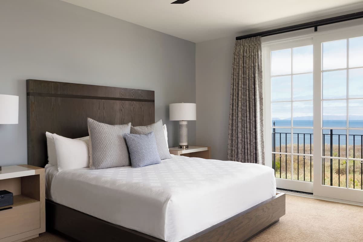 A hotel room with a large bed, two bedside tables with lamps, a view of the sea through a balcony, and a wooden headboard.