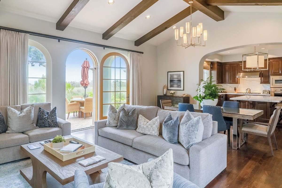A spacious living area with a grey sofa, a coffee table, and a dining table with chairs. There is a kitchen area with wooden cabinets and a stainless steel range hood. Large windows with curtains provide natural light and a view of the outdoors.
