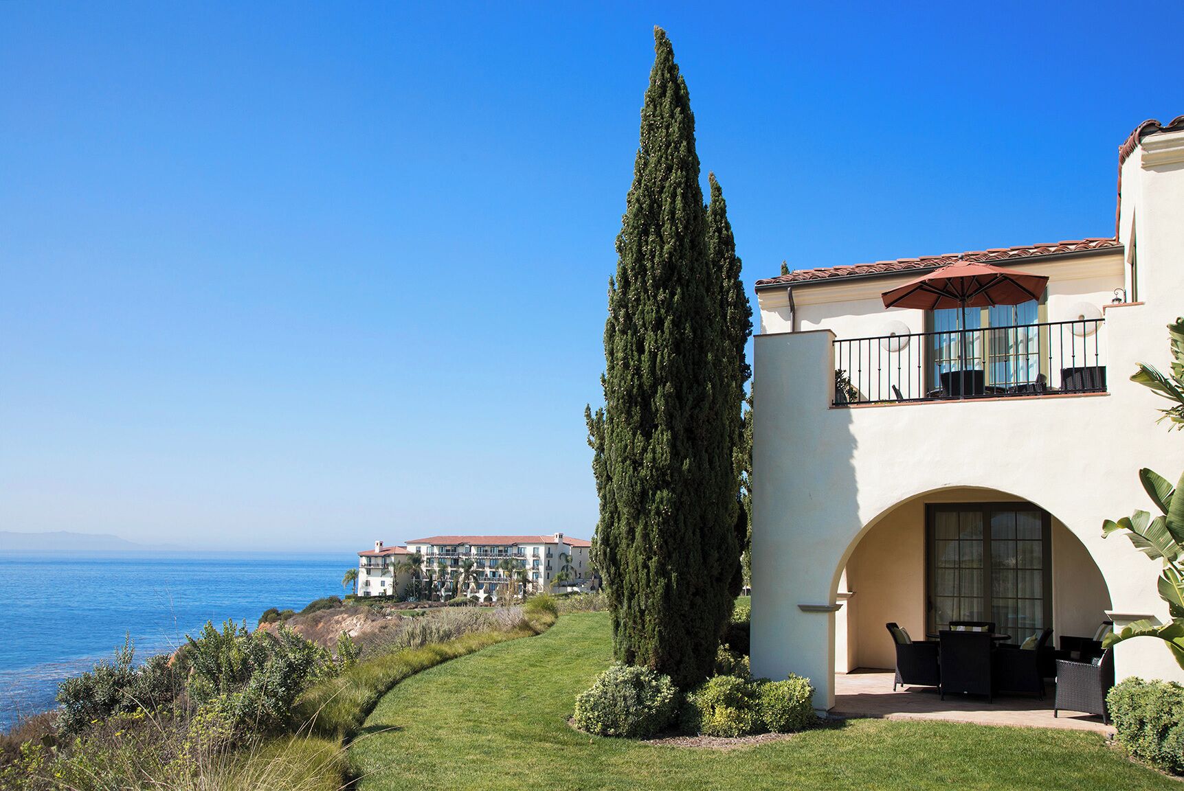 A coastal property with a large cypress tree, a balcony with outdoor furniture, and a view of the ocean.