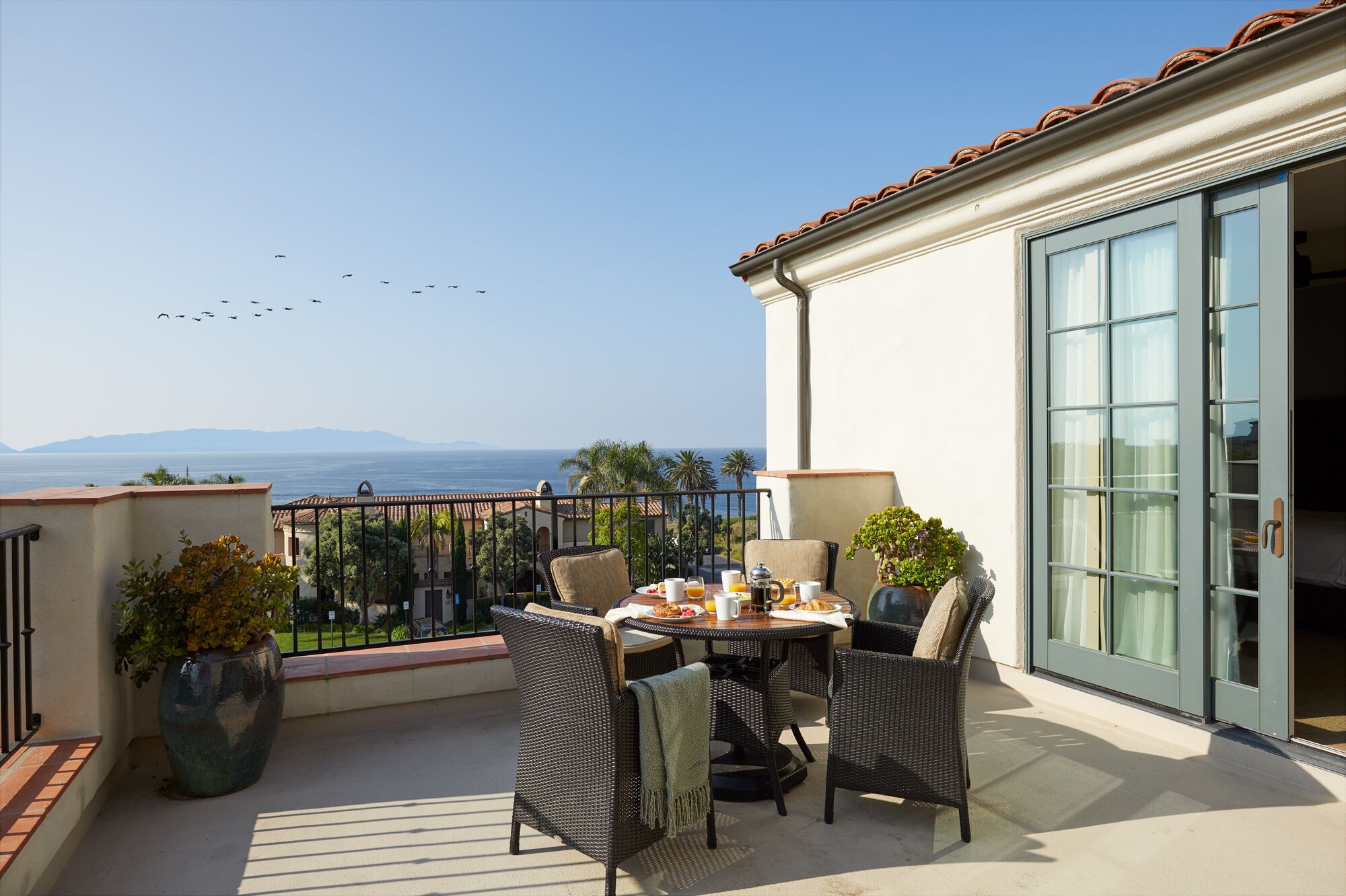 A balcony with wicker furniture, a table set for breakfast, and a view of the sea and mountains.