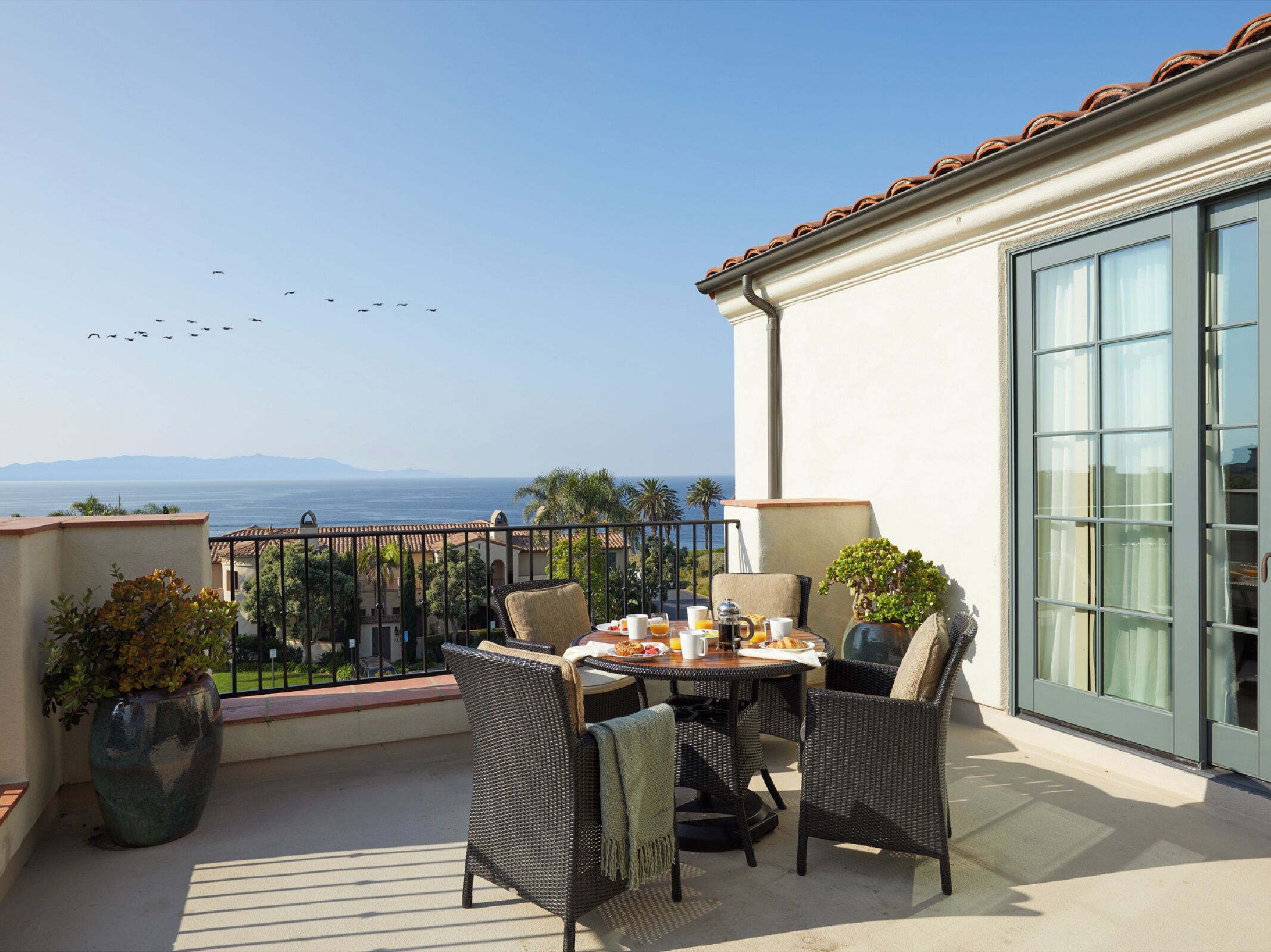 Un balcón con mesa lista para el desayuno, con vista al mar y aves volando en el cielo.