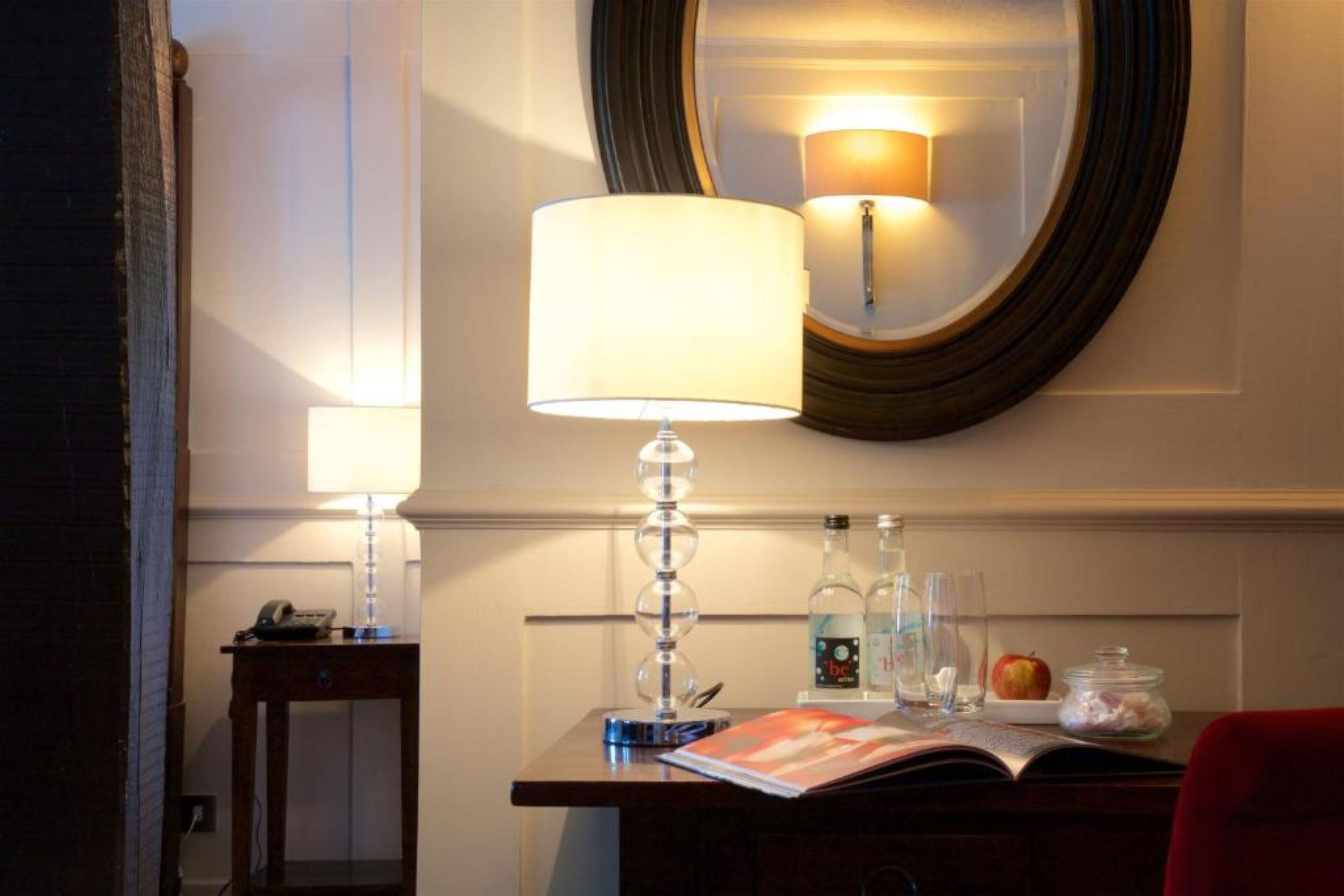 Une chambre d’hôtel équipée d’un bureau, d’une lampe, d’un miroir et d’une petite table sur laquelle se trouvent un téléphone, un livre et quelques bouteilles.