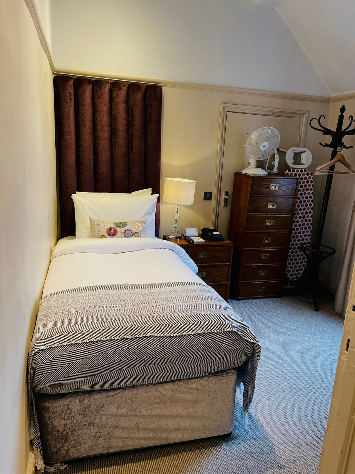 Una habitación de hotel con una cama, una mesita de noche, un tocador, una lámpara, un ventilador y un perchero.