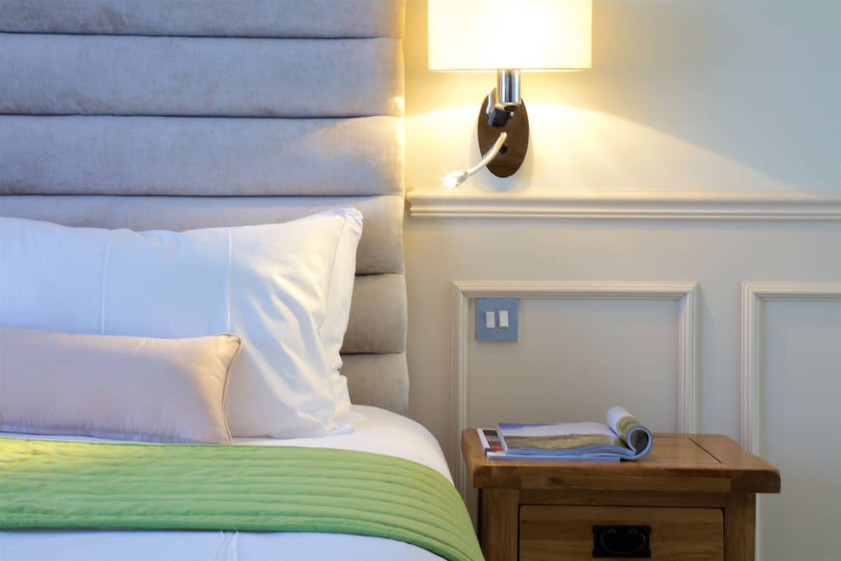 Una cama bien hecha con ropa de cama blanca y verde, una mesita de noche con una lámpara, y una lámpara de pared.