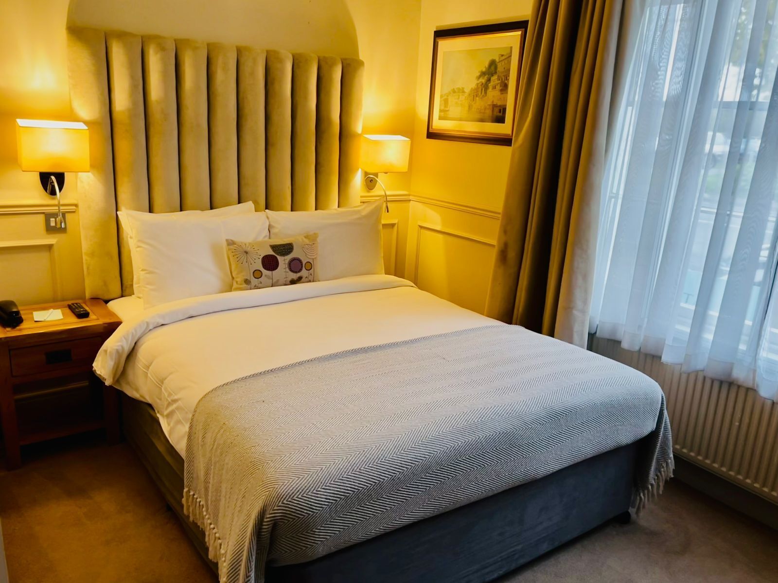 A hotel room with a large bed, two bedside lamps, a nightstand, and a window with sheer curtains.