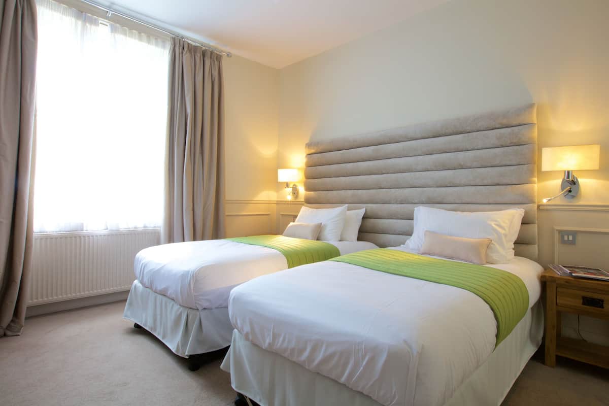 Habitación de hotel con dos camas, un ventanal con cortinas, un radiador y lámparas de noche.