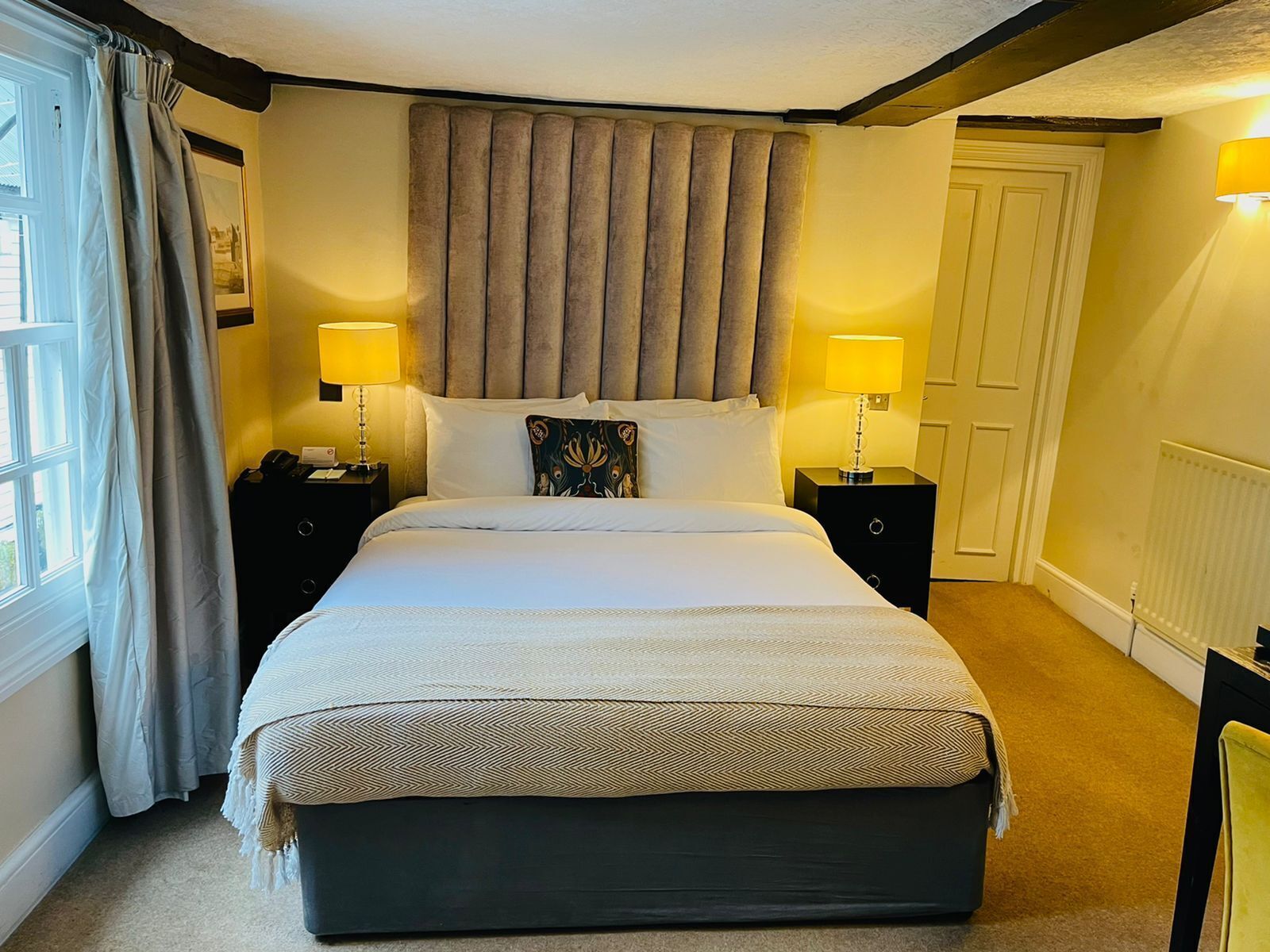 A neatly arranged bedroom with a large bed, two bedside tables with lamps, a window with curtains, and a door.