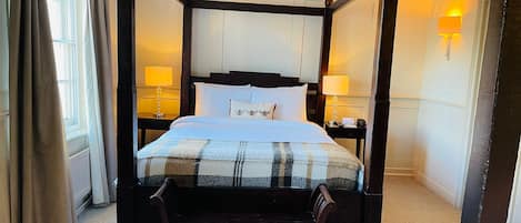 Una cama con dosel, con una colcha escocesa, dos mesitas de noche con lámparas, y un banco al pie de la cama.