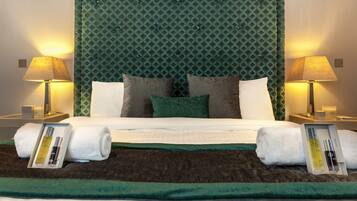 A neatly made bed with white linens, two pillows, and a green throw blanket. There are two bedside lamps with lampshades, a framed photo, and a small table with a candle and a box.