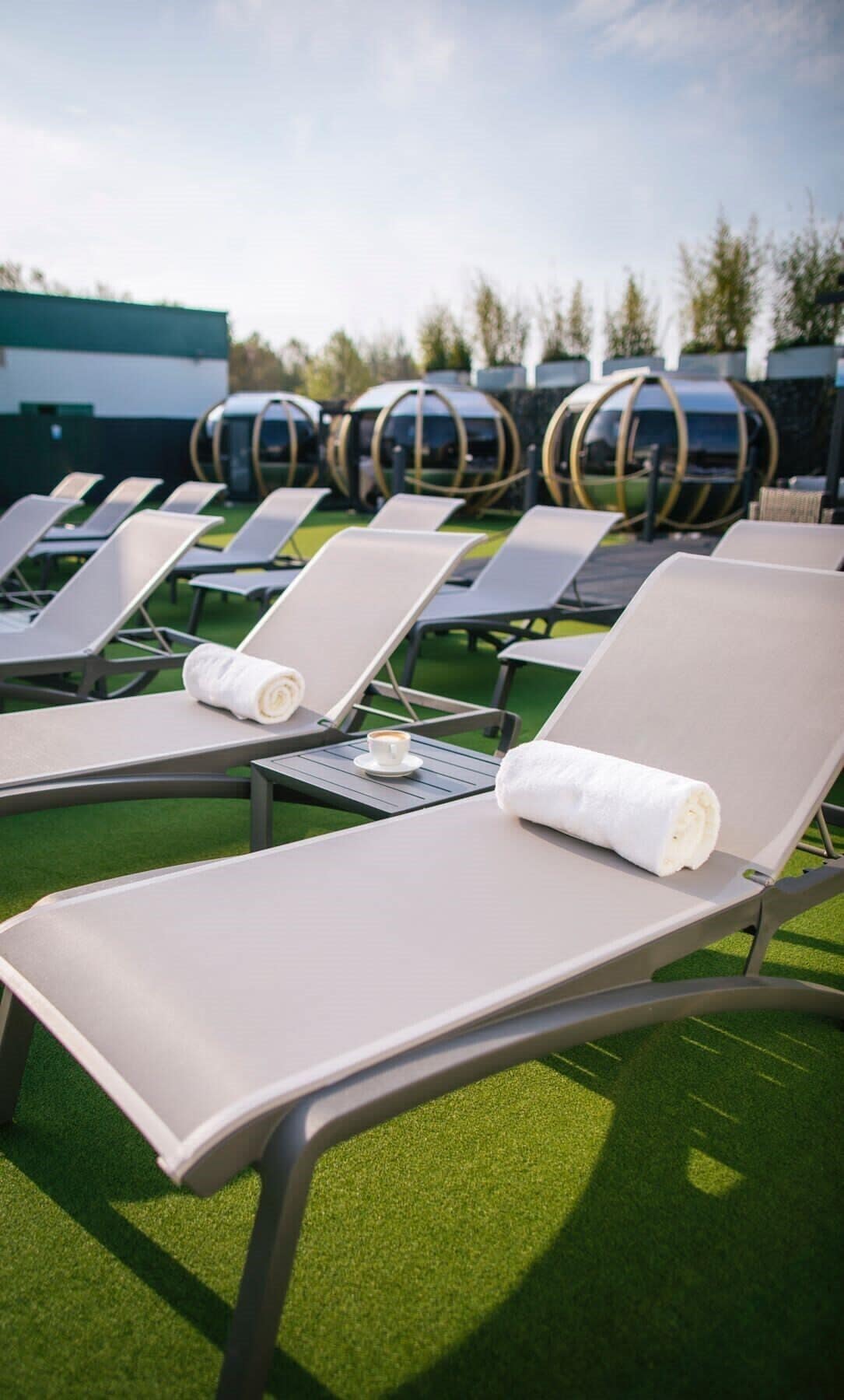 A row of sun loungers with towels and a cup of coffee on a table, set on artificial grass with a building in the background.