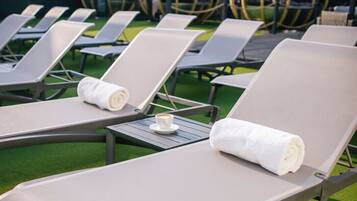 A row of sun loungers with towels and a cup of coffee on a table, set on artificial grass with a building in the background.
