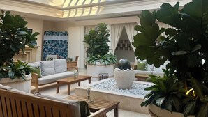 Un vaste espace intérieur doté d’un haut plafond et d’une verrière, comprenant un coin salon avec des canapés et une table basse, le tout agrémenté de grandes plantes en pot et d’un élément décoratif central en pierre.