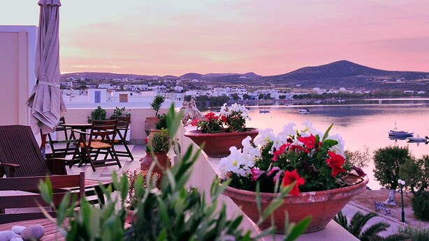 A terrace with outdoor seating, potted plants, and a view of a lake and distant buildings. - Portiani Hotel (Milos)